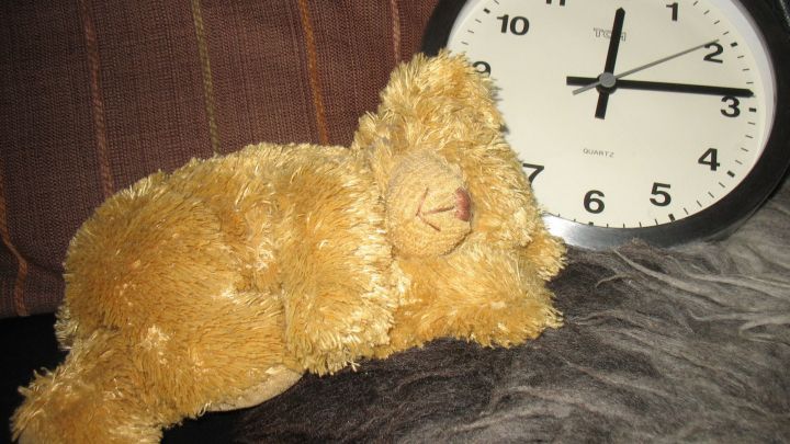 Ein pl&uuml;schiger L&ouml;we liegt entspannt neben einer Uhr auf einem weichen Untergrund, ideal f&uuml;r ein gesundes Mittagsschl&auml;fchen.