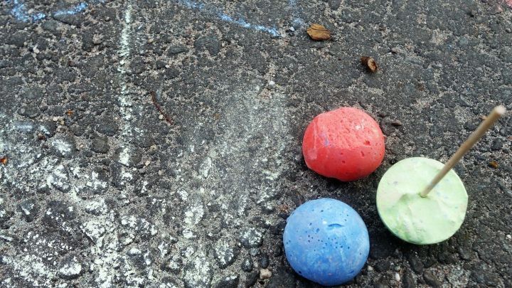 Bunte Stra&szlig;enmalkreide am Stiel liegt auf dem Asphalt neben einem blauen und roten Farbklecks und einer Zeichnung einer Blume.