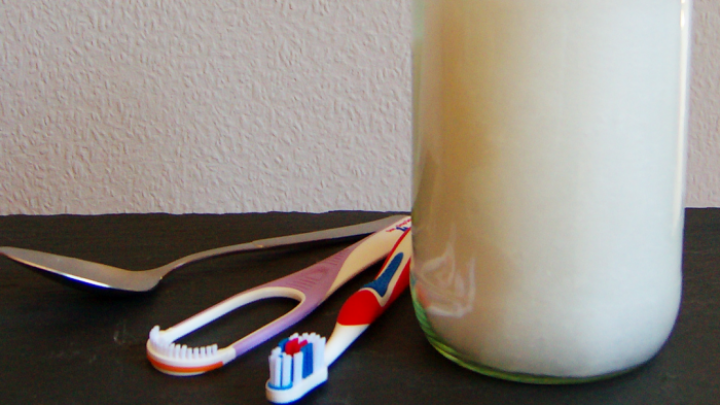 Ein Glas mit Kokos&ouml;l steht auf einem Tisch, neben einer Zahnb&uuml;rste und einem L&ouml;ffel, bereit f&uuml;r &Ouml;lziehen.