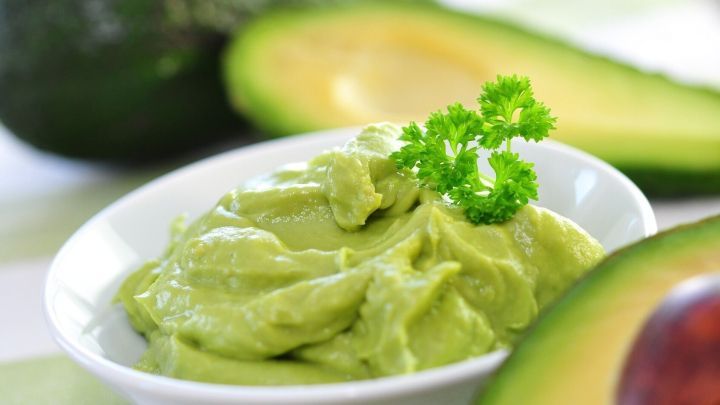 Ein cremiger Avocado-Dip liegt in einer wei&szlig;en Sch&uuml;ssel, umgeben von frischen Avocados auf einem hellen Tisch.