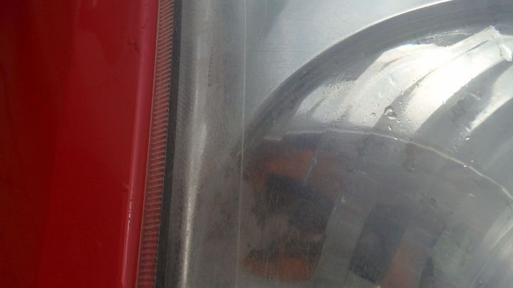 Tr&uuml;be Scheinwerfer einer roten Fahrzeugfront reflektieren schwaches Licht, umgeben von einer unscharfen Umgebung.