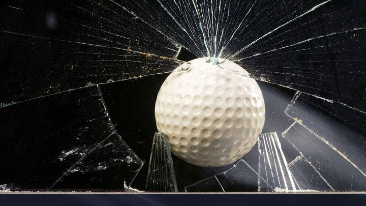 Ein Golfball trifft ein Glas, das dabei zerbricht. Der Moment zeigt den Aufprall und die Crack-Spuren im Glas.