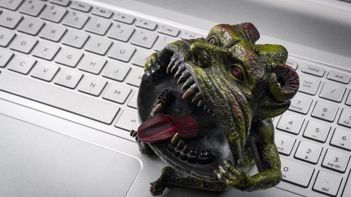Ein trollartiges Monster sitzt auf einer Laptop-Tastatur und zeigt seine Zunge, w&auml;hrend es drohend grinst.