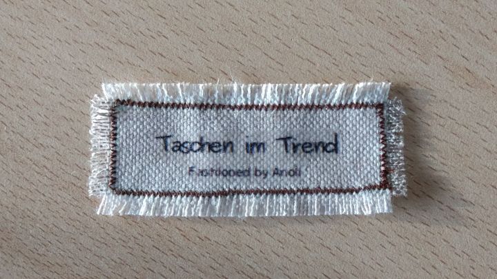 Ein ausgefranstes Stofflabel zeigt "Taschen im Trend" in stilvollem, minimalistischen Design auf hellem Hintergrund.