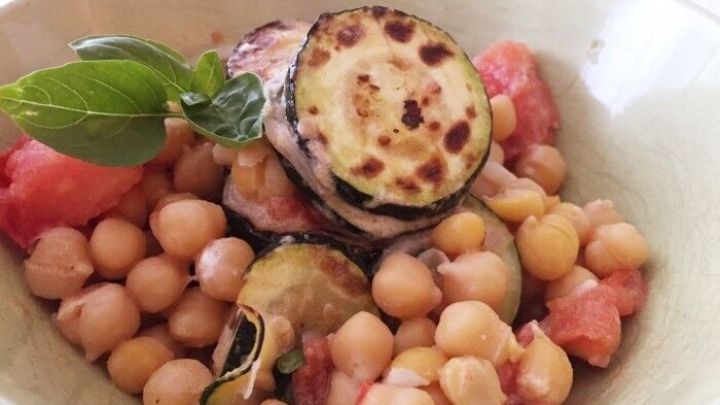 Ein bunter Kichererbsensalat mit gegrilltem Gem&uuml;se und frischen Kr&auml;utern in einer Schale, serviert auf einem Tisch.