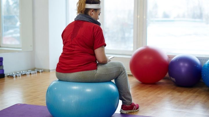 Eine Person sitzt auf einem blauen Gymnastikball in einem hellen Raum mit Fitnessger&auml;ten und bunten B&auml;llen im Hintergrund.