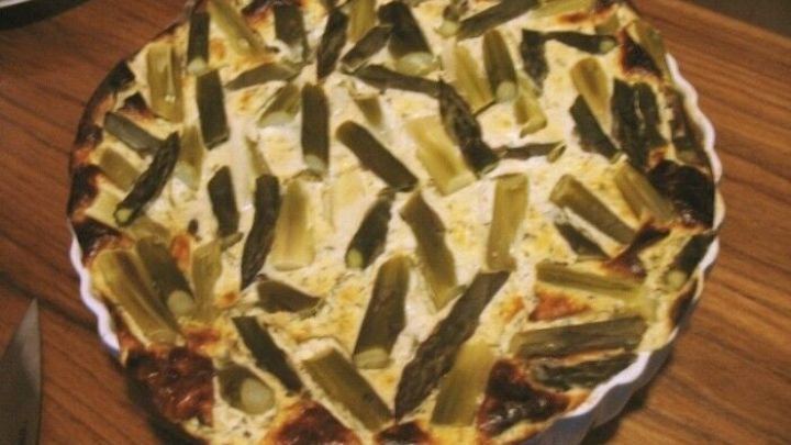 Eine K&auml;se-Spargel-Tarte liegt goldbraun auf einem Holzbrett, umgeben von einem rustikalen Ambiente in einer K&uuml;che.