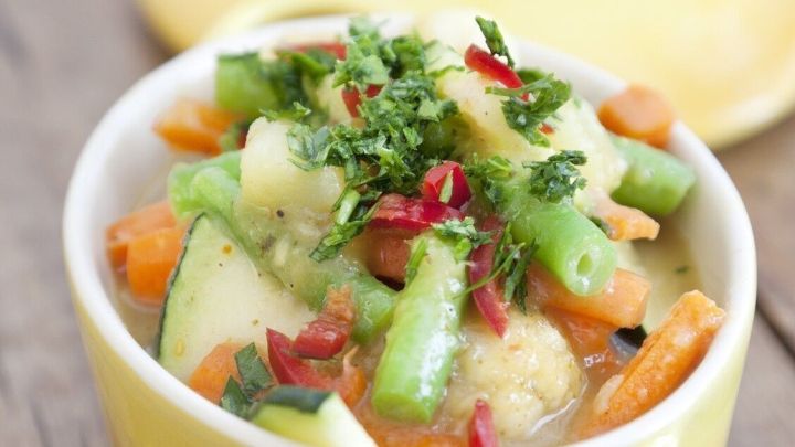 Ein gelbes Sch&auml;lchen mit einem bunten Fr&uuml;hlingseintopf aus Gem&uuml;se, frisch garniert und auf einem Holztisch serviert.