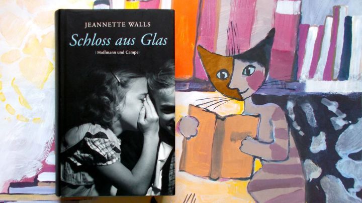 Ein Buch "Schloss aus Glas" von Jeannette Walls liegt auf einer bunten, k&uuml;nstlerisch gestalteten Unterlage, umgeben von B&uuml;cherstapeln und einer Katze.