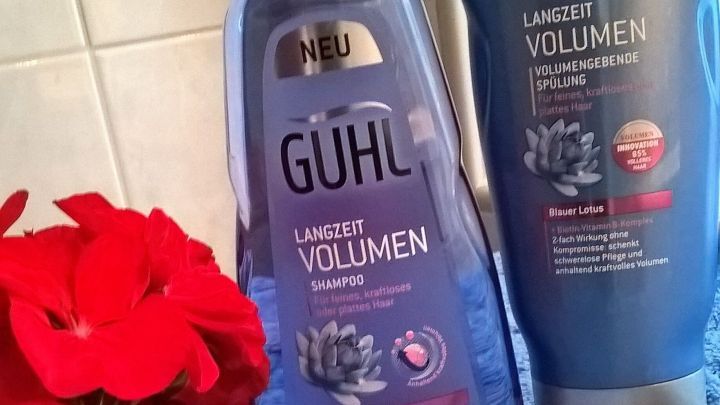 Shampoo und Sp&uuml;lung von Guhl f&uuml;r l&auml;ngeres Volumen stehen neben einer roten Blume auf einem blauen Handtuch im Badezimmer.