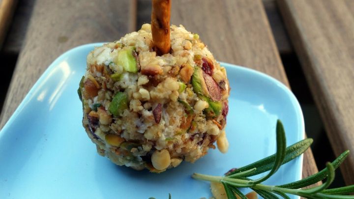 Eine K&auml;se-Pop-Kugel auf einem blauen Teller, umgeben von gehackten N&uuml;ssen und frischen Kr&auml;utern, auf einem Holztisch.