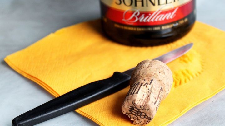 Eine Sektflasche steht auf einem gelben Tuch, w&auml;hrend ein Knife den Korken sanft zur&uuml;ck in die Flasche f&uuml;hrt.