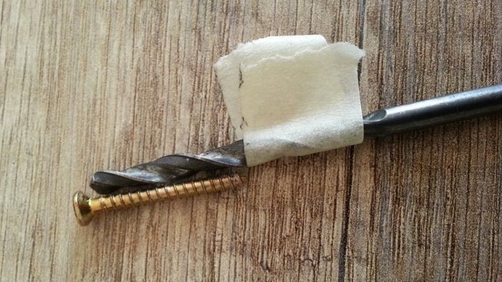 Ein Bohrer mit einem St&uuml;ck Klebeband ist auf einer Holzoberfl&auml;che platziert, daneben liegt eine Schraube.