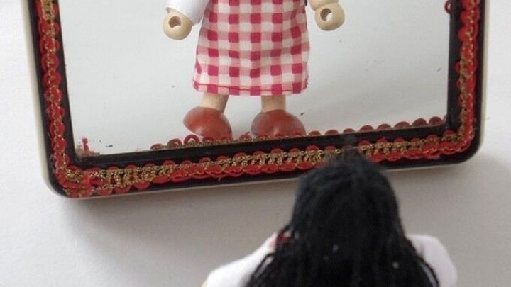 Eine Puppe mit langen, schwarzen Haaren sieht in einen Spiegel, der in einer hellen Umgebung steht, und betrachtet ihr Outfit.