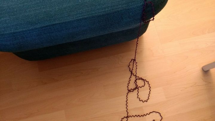Ein gestrandeter Wollfaden liegt entspannt auf einem Sofa und ist teilweise aufgeribbelt, umgeben von einem Holzfu&szlig;boden.