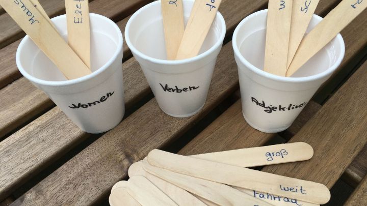 Drei Becher auf einem Tisch, gef&uuml;llt mit Holzst&auml;bchen, die W&ouml;rter wie "Verb", "Substantiv" und "Adjektiv" beschreiben.