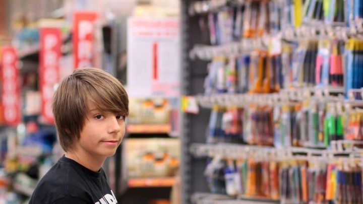 Ein Junge steht in einem Gesch&auml;ft f&uuml;r Schulbedarf und schaut auf Regale voller Artikel, w&auml;hrend er einen roten Einkaufswagen h&auml;lt.