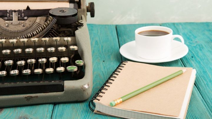 Eine Schreibmaschine steht auf einem blauen Tisch, w&auml;hrend eine Tasse Kaffee und ein Notizbuch daneben liegen.