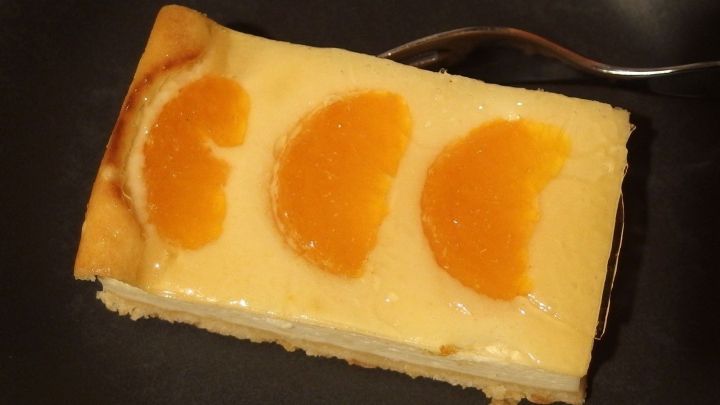 Ein St&uuml;ck K&auml;sekuchen mit orangefarbenen Fruchtscheiben liegt auf einem dunklen Teller, bereit zum Genie&szlig;en.