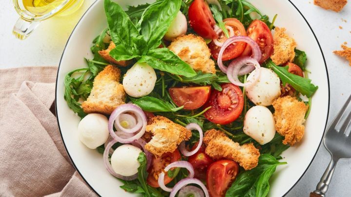 Ein bunter Knoblauch-Brotsalat mit frischen Tomaten, Mozzarella, Rucola und Basilikum in einer Sch&uuml;ssel, umgeben von Oliven&ouml;l.
