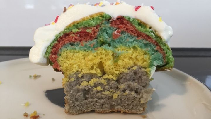 Ein bunter Muffin mit mehreren Schichten in Regenbogenfarben und einer wei&szlig;en Sahnehaube liegt auf einem Teller.