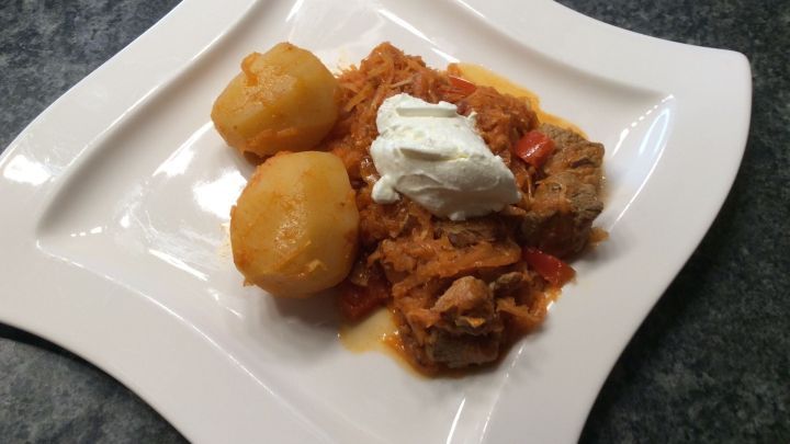 Auf einem wei&szlig;en Teller liegt Szegediner Krautfleisch, umgeben von Kartoffeln und einem Klecks Sauerrahm.