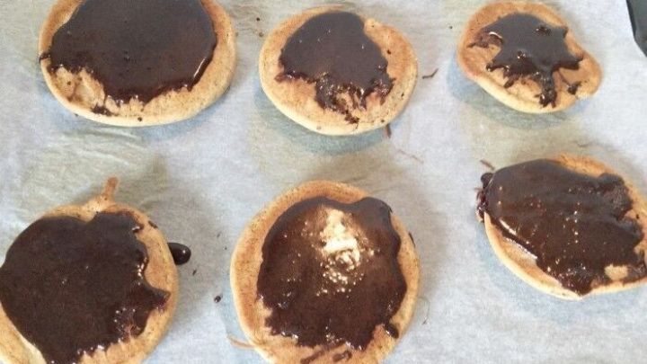 Sechs runde Kekse aus Buchweizenmehl liegen auf Backpapier, mit fl&uuml;ssiger Schokolade &uuml;berzogen, in einer K&uuml;che.