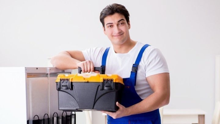 Ein Handwerker steht neben einem K&uuml;hlschrank und h&auml;lt eine Werkzeugkiste in der K&uuml;che, bereit f&uuml;r Reparaturen.