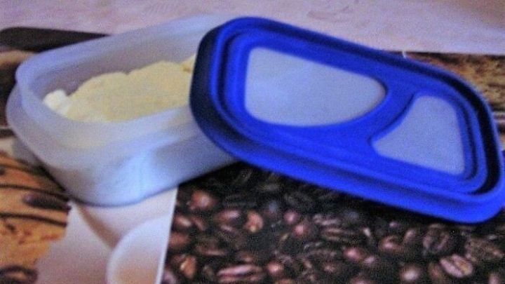 Eine geschlossene Plastikkonserve mit geriebenem Parmesan steht auf einem Tisch mit Kaffeebohnendekor. Der Deckel ist teilweise ge&ouml;ffnet.