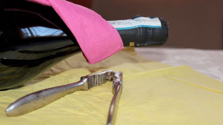 Eine Sektflasche mit pinkem Tuch liegt auf einem gelben Tisch, daneben liegt ein Nussknacker zum &Ouml;ffnen des Korkens.