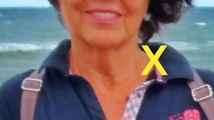 Eine Person im Vordergrund l&auml;chelt und zeigt den Halsbereich, w&auml;hrend im Hintergrund das Meer zu sehen ist.