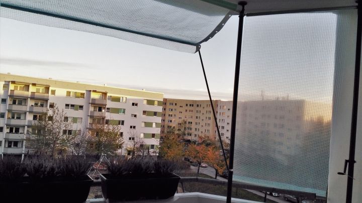 Eine Wintermarkise sch&uuml;tzt den Balkon vor N&auml;sse, w&auml;hrend im Hintergrund Wohnbl&ouml;cke und ein sanfter Sonnenuntergang sichtbar sind.