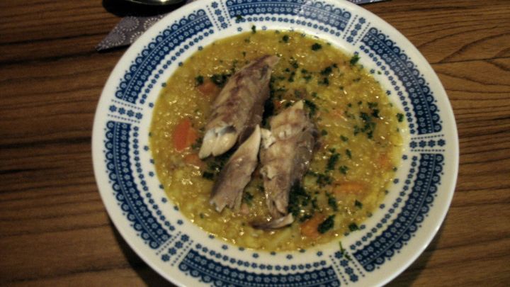 Ein Teller Linseneintopf mit ger&auml;uchertem Fischfilet liegt auf einem Holztisch, umgeben von Besteck und einer Serviette.