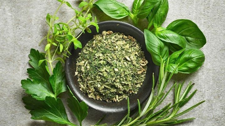 Ein schwarzes Sch&auml;lchen mit getrockneten Kr&auml;utern steht in der Mitte, umgeben von frischen Kr&auml;utern wie Basilikum und Rosmarin auf einer grauen Oberfl&auml;che.