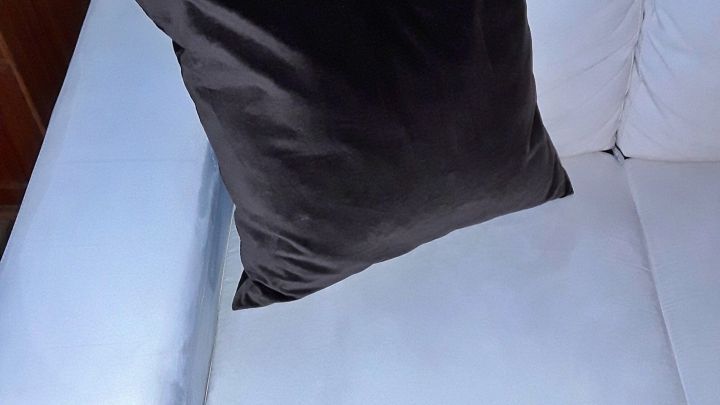 Ein helles Sofa mit einem gro&szlig;en, dunklen Kissen. Der Raum ist einfach und ordentlich gestaltet, sauber und einladend.