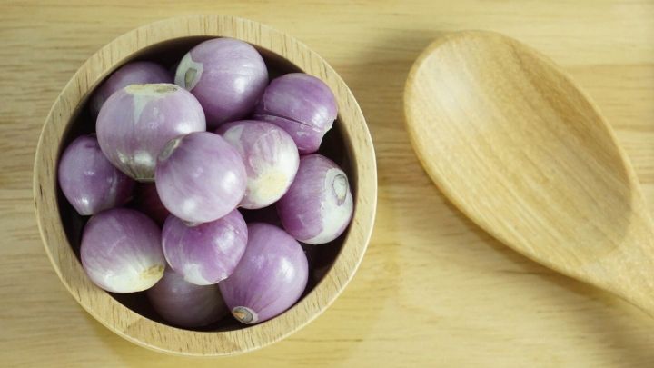 Eine Sch&uuml;ssel gef&uuml;llt mit kleinen lila Zwiebeln steht auf einem Holzuntergrund, daneben liegt ein Holzl&ouml;ffel.