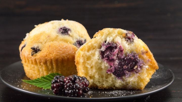 Zwei goldbraune Brombeer-Muffins liegen auf einem schwarzen Teller, umgeben von frischen Brombeeren und einem gr&uuml;nen Blatt.