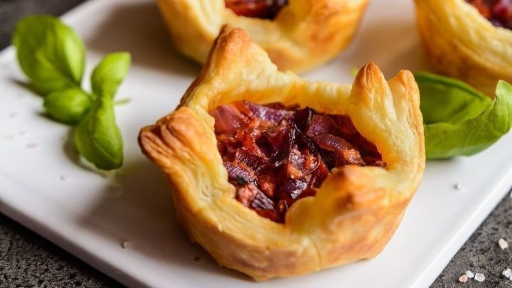 Kleine gef&uuml;llte H&auml;ppchen aus Bl&auml;tterteig liegen appetitlich auf einem wei&szlig;en Teller, umgeben von frischem Basilikum und roten Zwiebeln.