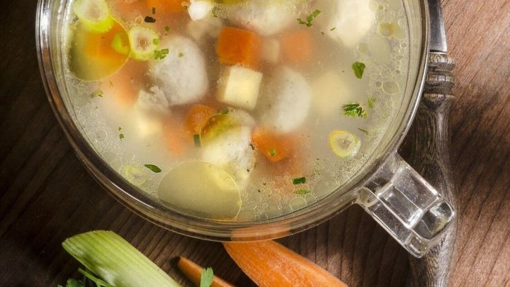 Eine klare Br&uuml;he mit buntem Gem&uuml;se und kleinen Einlagen in einem Glasbeh&auml;lter, auf einem Holztisch angerichtet.