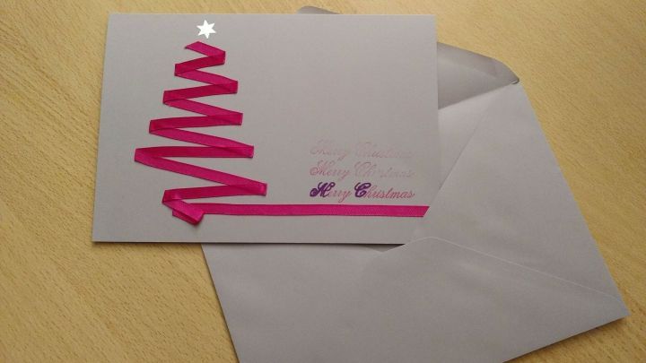Eine Weihnachtskarte mit einem bunten Band in Baumform liegt auf einem Holztisch neben einem grauen Umschlag.