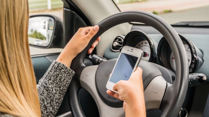 Eine Frau h&auml;lt ein Handy in der Hand, w&auml;hrend sie am Steuer sitzt, was im Stra&szlig;enverkehr gef&auml;hrlich ist.