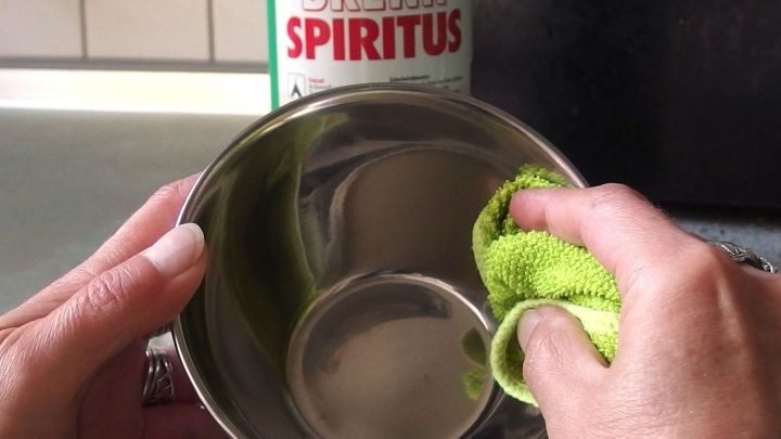 Eine Person reinigt eine gl&auml;nzende Edelstahlsch&uuml;ssel mit einem gr&uuml;nen Tuch, w&auml;hrend Brennspiritus im Hintergrund steht.