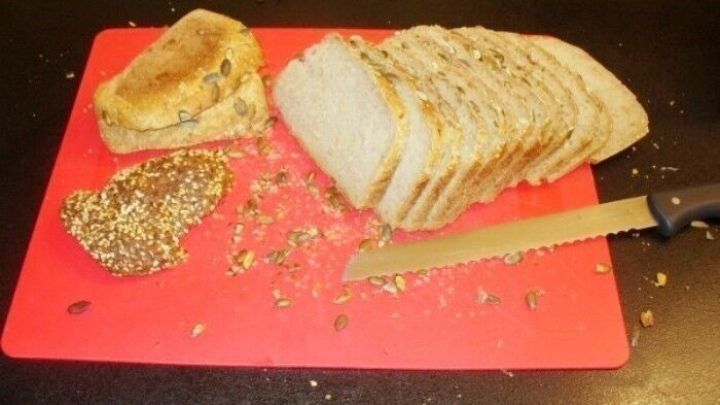 Ein frisch geschnittenes Brot mit Saaten liegt auf einem roten Schneidebrett, umgeben von Brotkr&uuml;meln und einem Messer.