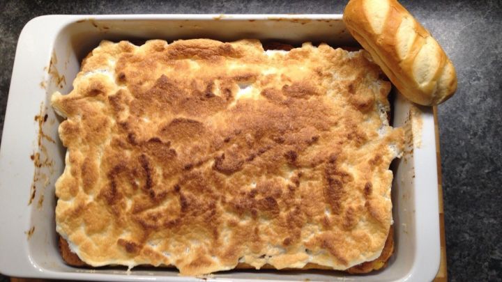 Ein Blech mit goldbraunem, gebackenem Auflauf aus Milchbr&ouml;tchen steht auf einem Tisch, daneben liegt ein frisches Br&ouml;tchen.