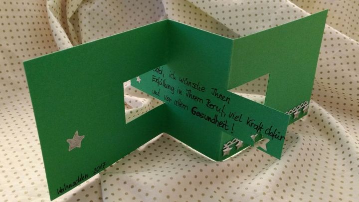 Eine aufgeschnittene Weihnachtskarte steht auf einer beige gestreiften Oberfl&auml;che, die in der Mitte gr&uuml;n ist und Sterne zeigt.