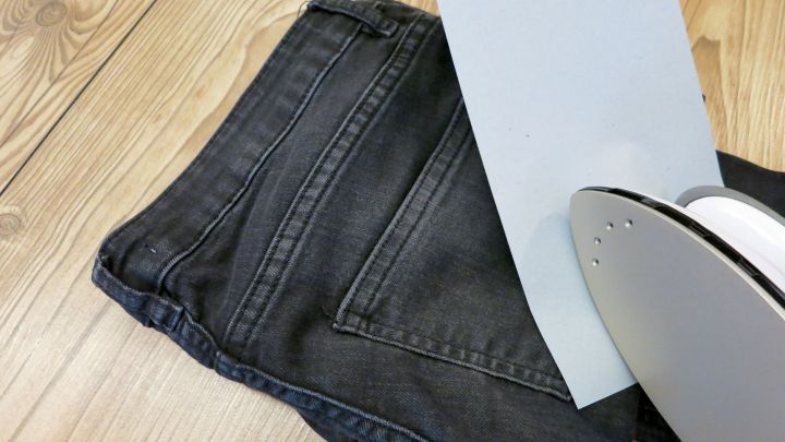 Auf einem Holzfu&szlig;boden liegen dunkle Jeans mit einem St&uuml;ck L&ouml;schpapier und einem B&uuml;geleisen daneben, bereit zum Entfernen von Wachsflecken.