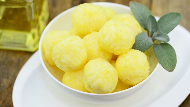 Kartoffelkl&ouml;&szlig;e liegen in einer Schale, mit frischen Bl&auml;ttern und einem Glas &Ouml;l im Hintergrund auf einem Holzbrett.