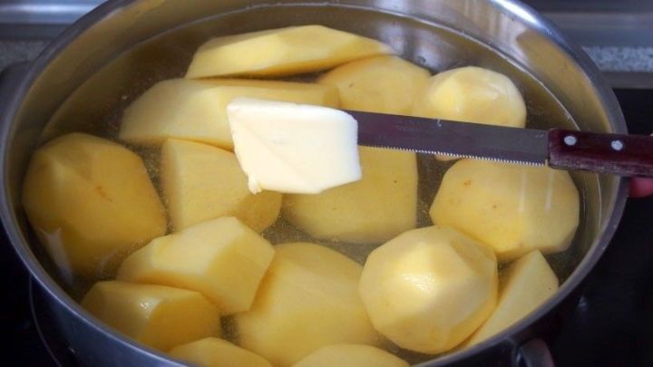Unsch&auml;lige Kartoffeln schwimmen in einem Topf mit Wasser, w&auml;hrend ein St&uuml;ck Butter mit einem Kochl&ouml;ffel hinzugef&uuml;gt wird.