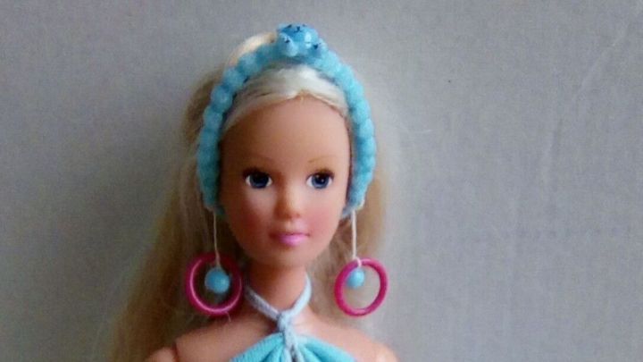 Eine Barbie-Puppe tr&auml;gt blaue Kleidung und gro&szlig;e Ohrringe, w&auml;hrend sie auf einem rosa Hocker sitzt.
