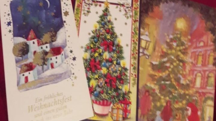 Drei festliche Weihnachtskarten liegen auf einem roten Untergrund und zeigen Illustrationen von einer Stadt, einem Weihnachtsbaum und einer winterlichen Szene.